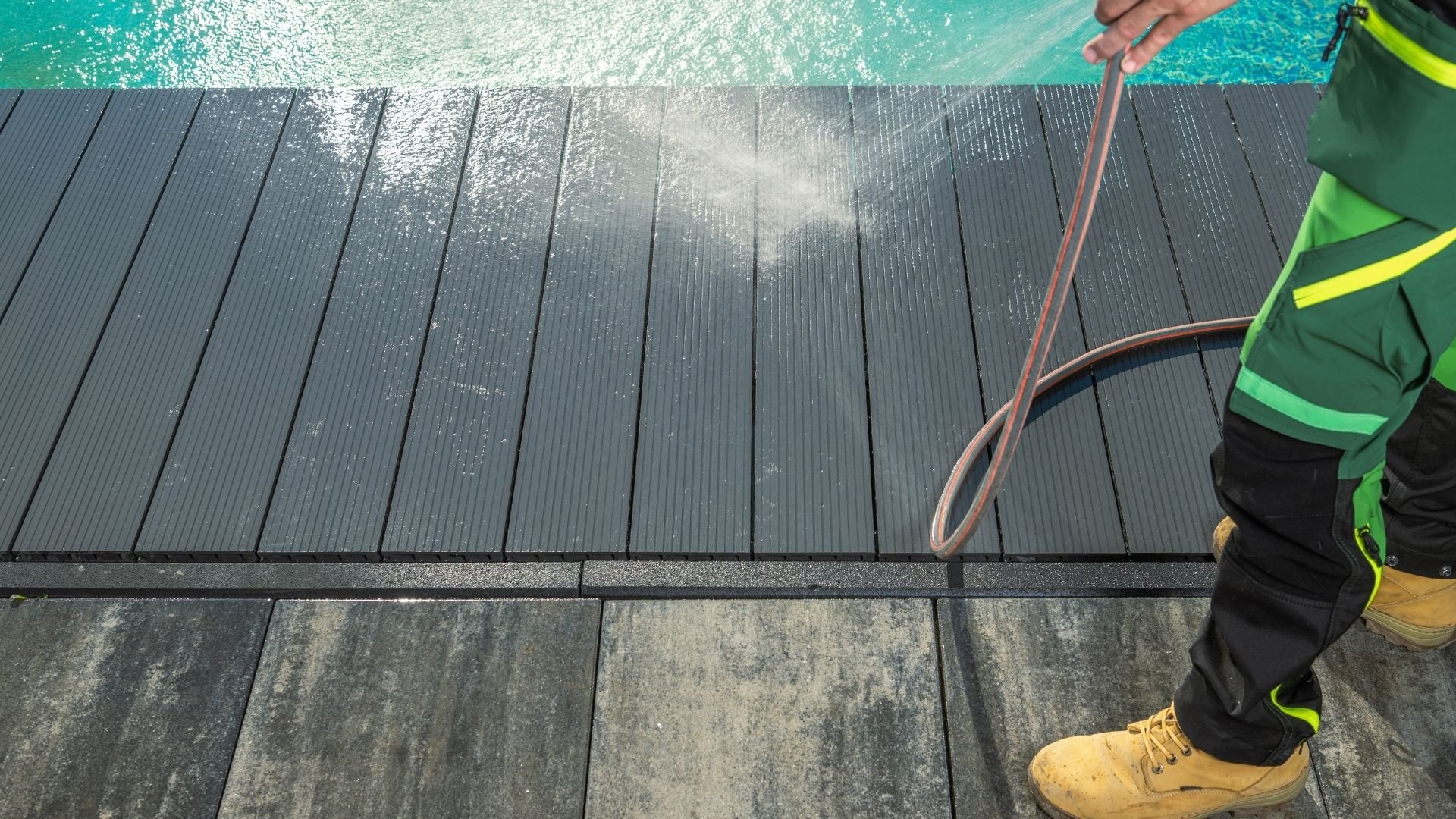 Worker spraying water on wooden deck beside turquoise ocean water.