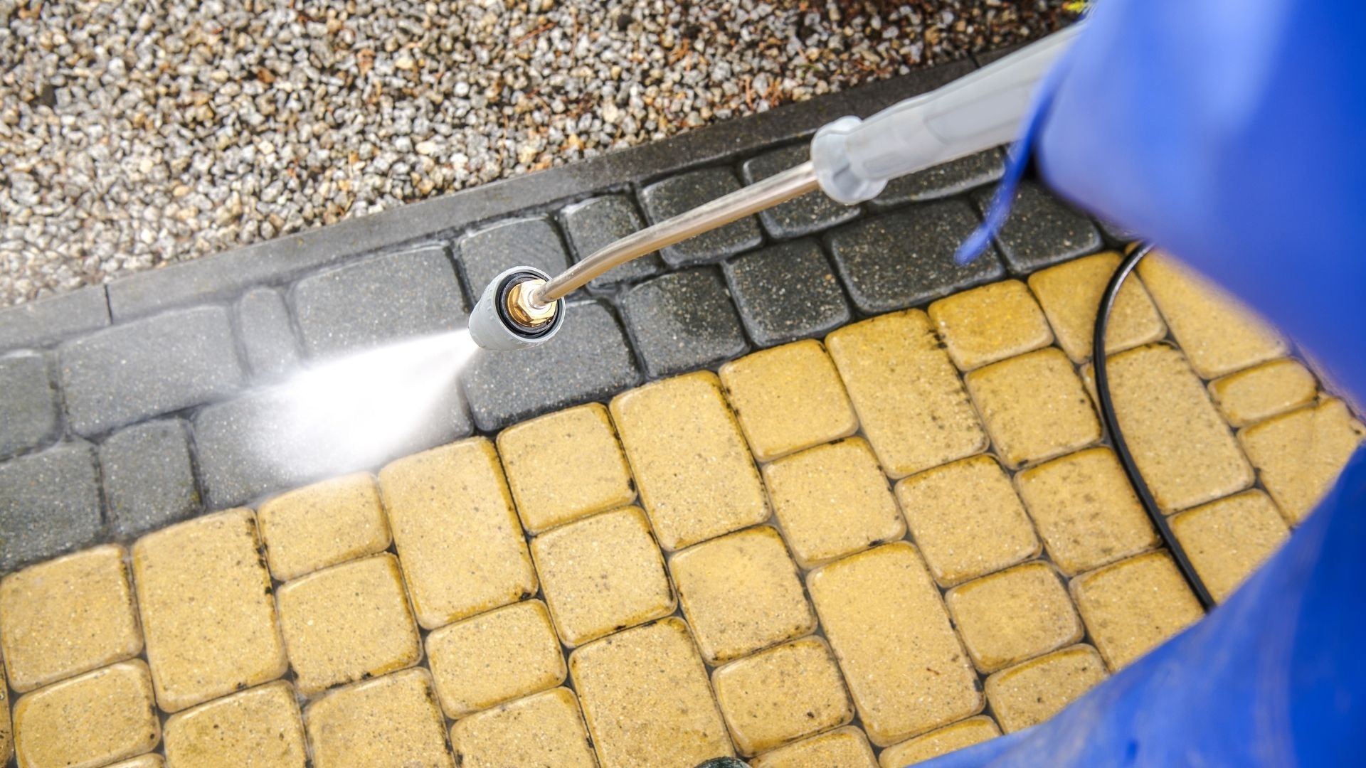 Pressure washer cleaning pavers between yellow and gray tiles with gravel above.